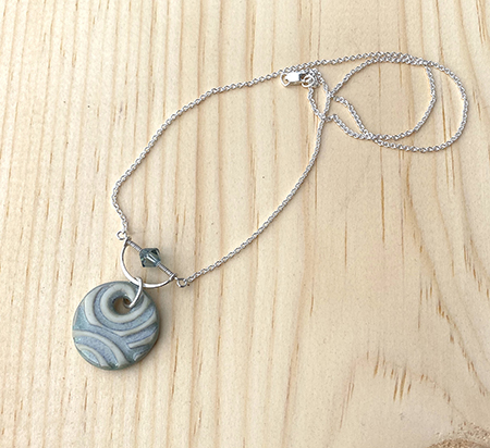 Swirly textured blue gray porcelain pendant with sterling silver half circle framing a matching crystal. The pendant is shown at an angle with chain gently coiled on a pine board background.